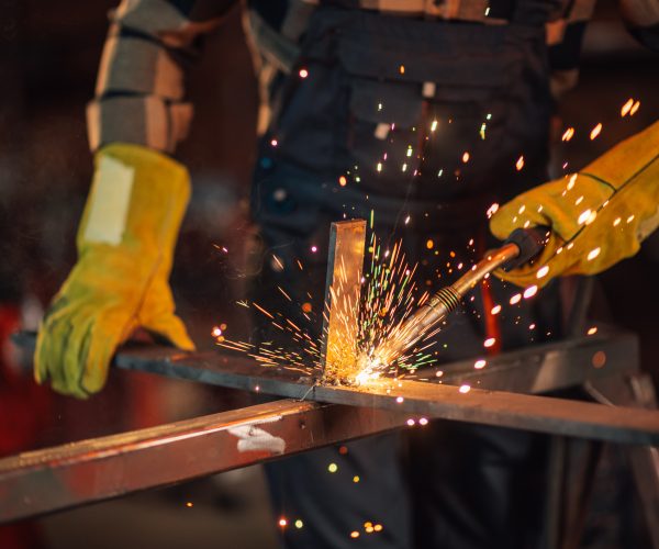Blurred background of a worker in a work overall wearing a safety equipment and protective gloves while welding a metal construction. Glowing sparks flying all around the working facility.Copy space.