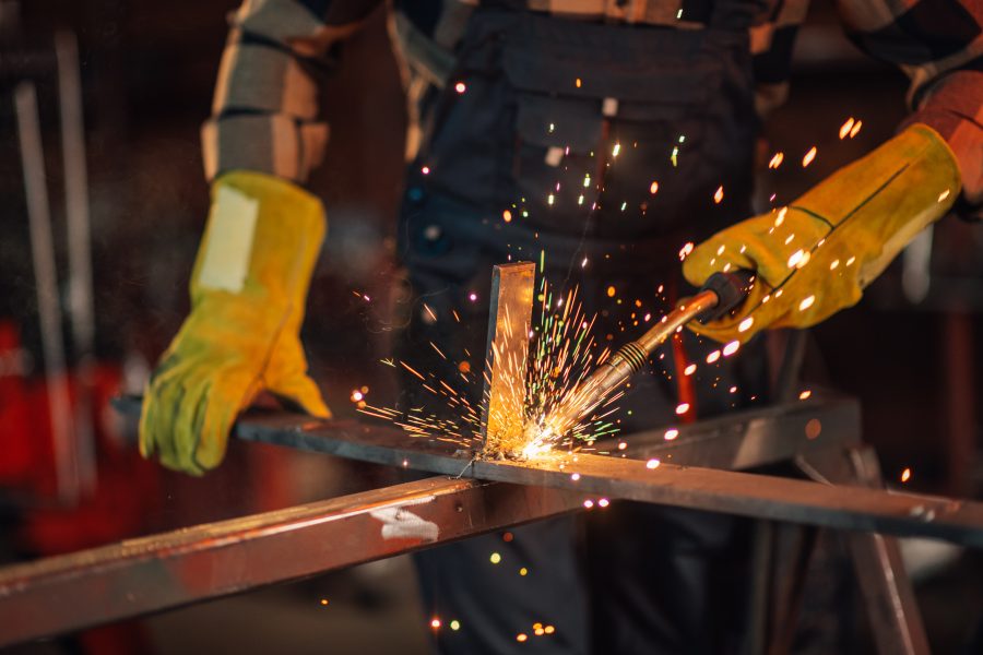 Blurred background of a worker in a work overall wearing a safety equipment and protective gloves while welding a metal construction. Glowing sparks flying all around the working facility.Copy space.